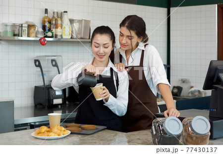 Young asian barista with a smile pouring milk from the jug into a paper coffee cup. Caucasian assistant in an apron stands watching beside her. Morning atmosphere in a coffee shop. Young asian barista with a smile pouring milk from the jug into a paper coffee cup. Caucasian assistant in an apron stands watching beside her. Morning atmosphere in a coffee shop. 77430137