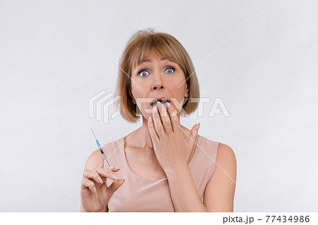 Pretty mature lady covering her mouth in shock, holding syringe with beauty injection on light studio background Pretty mature lady covering her mouth in shock, holding syringe with beauty injection on light studio background 77434986