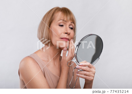 Portrait of mature woman checking her wrinkles in miror, looking at signs of aging on her face over light background 77435214