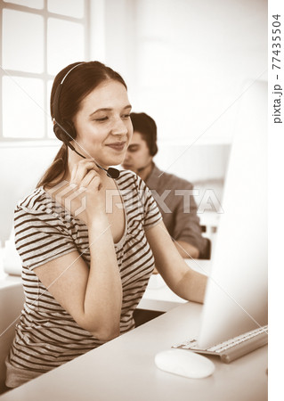 Casual dressed young woman using headset and computer while talking with customers online. Group of operators at work. Call center, business concept 77435504