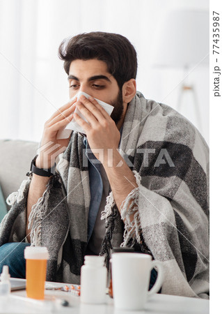 Stay home if feel sick. Ill arab guy covered in blanket sneezing his nose, sitting on sofa in living room, cropped Stay home if feel sick. Ill arab guy covered in blanket sneezing his nose, sitting on sofa in living room, cropped 77435987