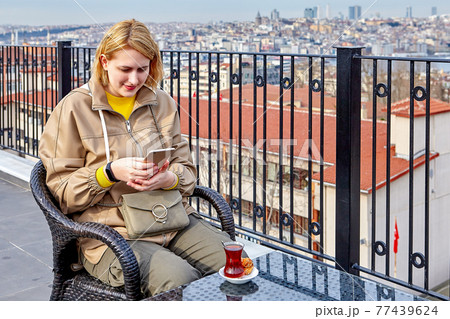 Tea Party on rooftop of hotel overlooking Istanbul cityscape, young woman scans online messages as hot drink cools. 77439624