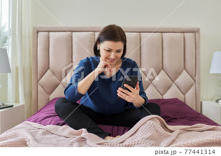 Smiling middle aged woman wearing wireless headphones looking at smartphone 77441314