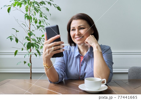 Mature woman with smartphone sitting at home smiling looking at phone screen Mature woman with smartphone sitting at home smiling looking at phone screen 77441665
