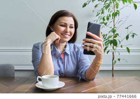 Mature woman with smartphone sitting at home smiling looking at phone screen Mature woman with smartphone sitting at home smiling looking at phone screen 77441666
