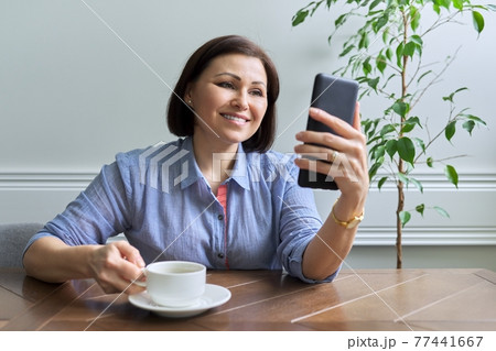 Mature woman with smartphone sitting at home smiling looking at phone screen Mature woman with smartphone sitting at home smiling looking at phone screen 77441667
