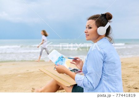 Mature woman sitting on sandy beach drawing sketch of sea Mature woman sitting on sandy beach drawing sketch of sea 77441819