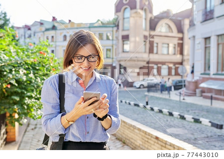 Mature business woman typing on smartphone walking along city street. 77443010