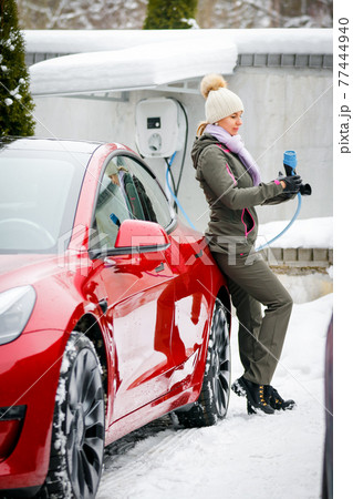Stylish girl stands near her red electric car and waits for the car to charge. Connecting the plug of the electric vehicle charger. Stylish girl stands near her red electric car and waits for the car to charge. Connecting the plug of the electric vehicle charger. 77444940
