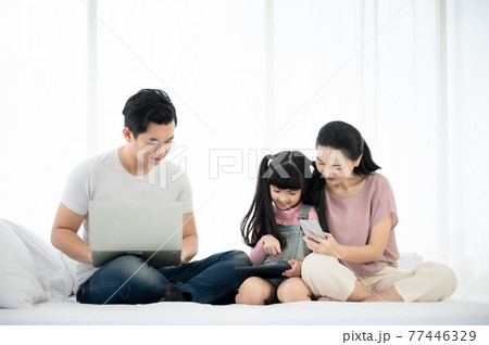 Father and mother with daughter relaxing and using laptop and smartphone on bed 77446329