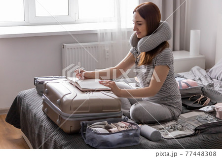 Smiling tourist woman packing suitcase to vacation writing paper list getting ready to travel trip Smiling tourist woman packing suitcase to vacation writing paper list getting ready to travel trip 77448308