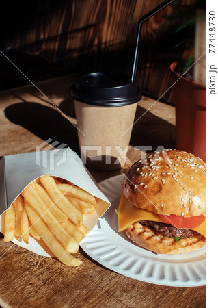 Burger in paper packaging, coffee and fries close-up. Food in a fast food restaurant. Macro photo of a burger with meat and vegetables, French fries and coffee. Fast food set Burger in paper packaging, coffee and fries close-up. Food in a fast food restaurant. Macro photo of a burger with meat and vegetables, French fries and coffee. Fast food set 77448730