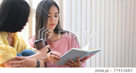 Photo of young sisters relaxing/reading a notebook while sitting together at the wooden floor over comfortable living room windows as background. 77451665