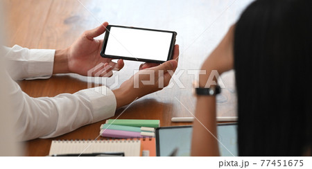 Photo of young businessman showing a white blank screen smartphone in his hands to colleague while sitting together at the wooden working table over orderly workspace as background. Photo of young businessman showing a white blank screen smartphone in his hands to colleague while sitting together at the wooden working table over orderly workspace as background. 77451675