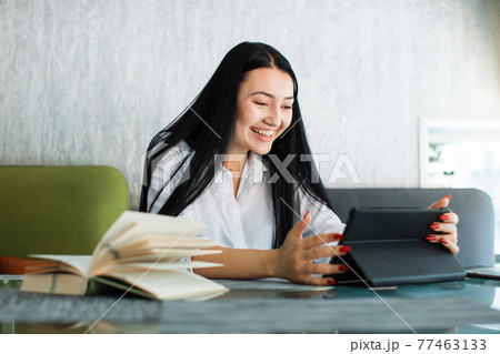Happy joyful likable Asian woman uses digital tablet, while sitting at the table in coffee shop. Young businesswoman having video call, smiling cheerfully to webcamera. 77463133