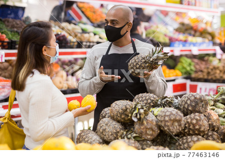 Grocery supermarket seller in protective mask helping woman choose delicious pineapple Grocery supermarket seller in protective mask helping woman choose delicious pineapple 77463146