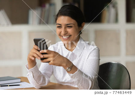 Happy Indian businesswoman looking at smartphone screen, sitting at desk Happy Indian businesswoman looking at smartphone screen, sitting at desk 77463449