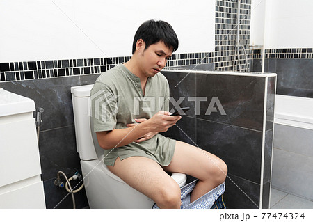 unhappy man with stomachache using smartphone and sitting in toilet bowl in bathroom at home 77474324