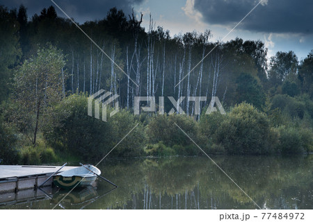 Water surface of the lake before a thunderstorm. Trees and clouds are reflected in the lake. A lone boat is moored to the dock. 77484972
