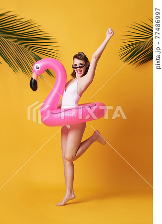 Happy young woman jumping on yellow background dressed in swimwear holding flamingo rubber ring beach. 77486997