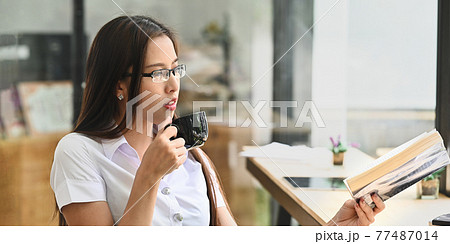 Brunette woman in glasses holding a coffee cup while relaxing/reading a book at the long wooden counter bar over comfortable restaurant as background. Brunette woman in glasses holding a coffee cup while relaxing/reading a book at the long wooden counter bar over comfortable restaurant as background. 77487014