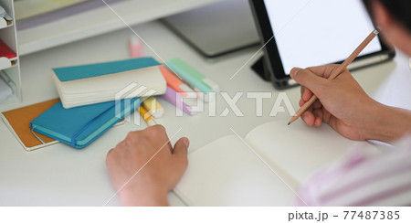 Cropped image of young student hands writing on notebook while sitting in front of white blank screen computer tablet that putting on white student desk. Student doing homework at home concept. 77487385