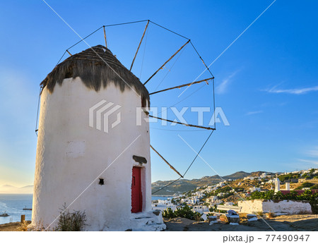 Famous tourist attraction of Mykonos, Greece. Traditional whitewashed windmill on hilltop. Summer, sundown, blue sky. Travel destination, iconic view. Famous tourist attraction of Mykonos, Greece. Traditional whitewashed windmill on hilltop. Summer, sundown, blue sky. Travel destination, iconic view. 77490947
