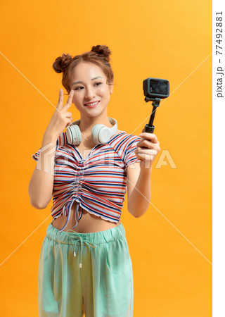 Woman capturing herself with small personal camera on yellow background 77492881