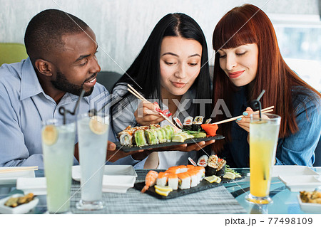 Two girls, Caucasian and Asian, and African guy, have a dinner with japanese sushi rolls in cafe, using food chopsticks. Close up portrait. Food, drink, asian meal, seafood and friendship concept. Two girls, Caucasian and Asian, and African guy, have a dinner with japanese sushi rolls in cafe, using food chopsticks. Close up portrait. Food, drink, asian meal, seafood and friendship concept. 77498109