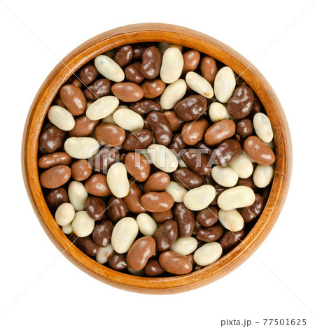 Roasted soybeans, coated with chocolate, in a wooden bowl. Snack of crispy roasted soybeans, covered with dark, milk and white chocolate. Close-up, from above, on white background, macro food photo. 77501625