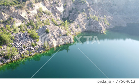 Rocky shore of the Radon Lake on a sunny summer morning. Aerial view of an old flooded granite quarry. A picturesque pond. Rocky shore of the Radon Lake on a sunny summer morning. Aerial view of an old flooded granite quarry. A picturesque pond. 77504612