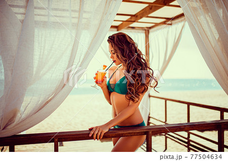 Portrait of sexy young woman in bikini with orange cocktail glass with straw and fruit posing at wooden bar of tent with white curtains. 77504634