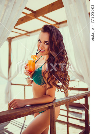 Portrait of sexy young woman in bikini with orange cocktail glass with straw and fruit posing at wooden bar of tent with white curtains. Portrait of sexy young woman in bikini with orange cocktail glass with straw and fruit posing at wooden bar of tent with white curtains. 77505033