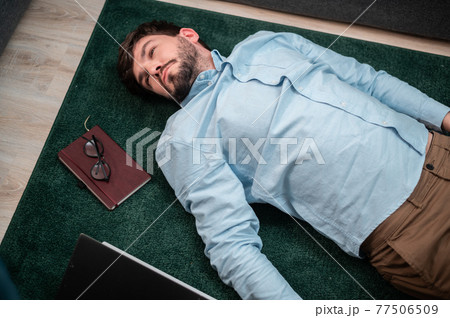 Young cute office worker lies on the floor mat next to the sofa, he is tired and asleep, top view 77506509