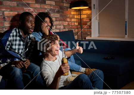 Emotional students watching sport match at home sitting on sofa. fans cheering for favourite national team, drinking beer Emotional students watching sport match at home sitting on sofa. fans cheering for favourite national team, drinking beer 77507380