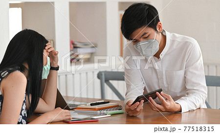 Photo of young couple that wearing a medical mask sitting and relaxing together at the wooden working desk over comfortable living room as background. 77518315