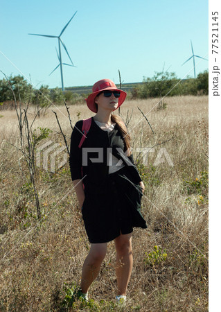 Woman in red hat in the field with wild grass blue sky wind turbines background. Stylish girl long brunette hair 77521145