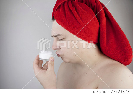Funny man with a red towel on his head sniffs, tastes and smears a cream on his face. The concept of male self-care, spa treatments and male cosmetics 77523373
