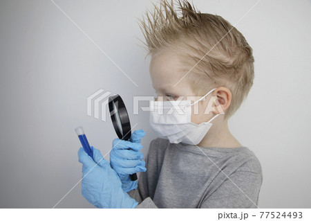 The child plays a doctor and a scientist. A boy in a medical mask studies a medicine in a test tube through a magnifying glass. The concept of education and promotion of medical professions 77524493