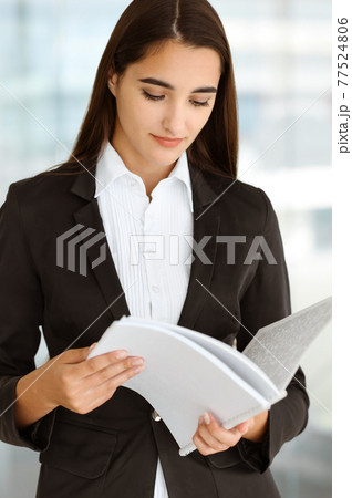 A friendly young dark hair businesswoman or female student dressed in a black suit is standing and looking through some papers in the office A friendly young dark hair businesswoman or female student dressed in a black suit is standing and looking through some papers in the office 77524806