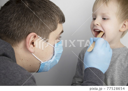 An otolaryngologist examines a child's throat with a wooden spatula. A possible diagnosis is inflammation of the pharynx, tonsils or pharyngitis. The concept of treatment and prevention of childhood An otolaryngologist examines a child's throat with a wooden spatula. A possible diagnosis is inflammation of the pharynx, tonsils or pharyngitis. The concept of treatment and prevention of childhood 77528654