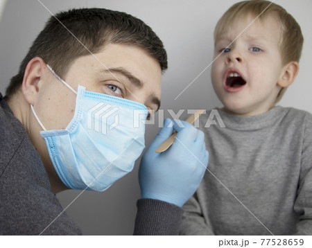 An otolaryngologist examines a child's throat with a wooden spatula. A possible diagnosis is inflammation of the pharynx, tonsils or pharyngitis. The concept of treatment and prevention of childhood An otolaryngologist examines a child's throat with a wooden spatula. A possible diagnosis is inflammation of the pharynx, tonsils or pharyngitis. The concept of treatment and prevention of childhood 77528659