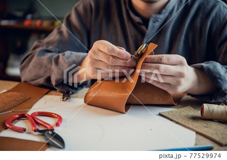 A young experienced shoemaker or tailor sews a unique piece of leather in a spacious bright studio 77529974