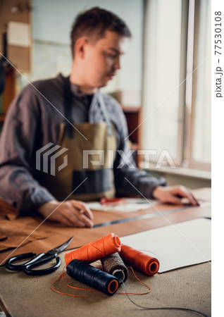 Several spools of colored thread on the desktop of a tailor, who is in the background in out-of-focus sewing a leather product 77530108