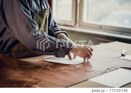A young apprentice transfers a pattern from paper to leather using a pencil in the workshop on the table 77530146