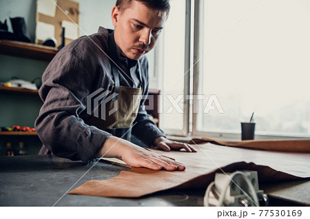 A young apprentice in a boot workshop prepares leather for further use on a large table A young apprentice in a boot workshop prepares leather for further use on a large table 77530169