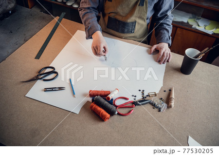 A young shoemaker makes a drawing for a pattern for leather shoes on a table in his workshop A young shoemaker makes a drawing for a pattern for leather shoes on a table in his workshop 77530205