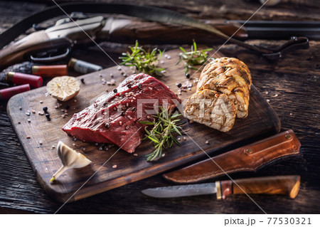 Nutrient rich raw deer venison prepared for a cooking process on a rustic wooden desk with roasted garlic, rosemary and huntig accesories like shot gun and ammunition Nutrient rich raw deer venison prepared for a cooking process on a rustic wooden desk with roasted garlic, rosemary and huntig accesories like shot gun and ammunition 77530321