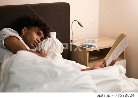 Young guy lying in the bed at home, resting and reading an interesting book before going to sleep 77533024