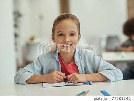 Lovely pupil. Gorgeous little school girl smiling at camera while sitting at the desk in classroom 77533246